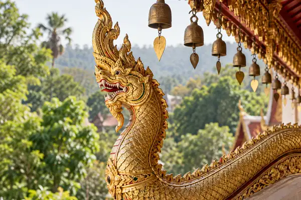 Gold naga serpent railing and small bells on a temple eave with green trees behind